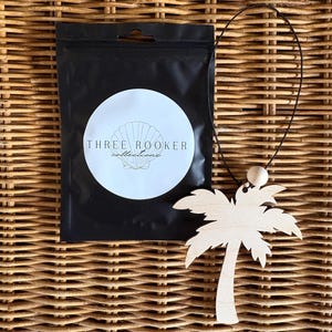 May include: A wooden palm tree pendant with a black cord and a small wooden bead. The pendant is light brown and is displayed on a woven wicker surface. A black pouch with the text "THREE ROOKER collections" is also in the image.