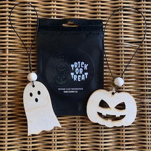 May include: Two light-colored wooden car fresheners, one shaped like a ghost and one like a jack-o'-lantern, each with a black elastic cord and a white bead. They are displayed in front of a black pouch labeled Trick or Treat Wood Car Freshener.