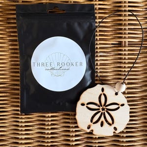 May include: A light-colored wooden sand dollar ornament featuring a laser-cut floral pattern, attached to a black cord with a small wooden bead. Next to it is a black plastic pouch with a white circular label displaying the text THREE ROOKER collections.