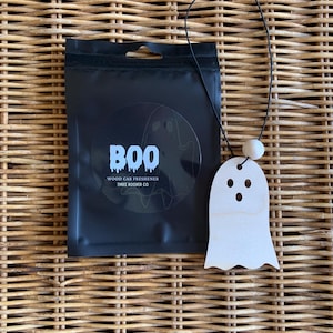 May include: A black rectangular pouch with the text BOO WOOD CAR FRESHENER THREE ROOKER CO and a ghost graphic, paired with a light-colored wooden ghost-shaped ornament hanging from a black string with a small round wooden bead.