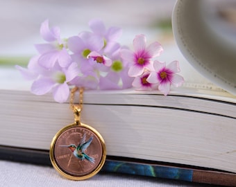 May include: A gold-colored pendant necklace featuring a copper coin with a painted hummingbird design. The pendant is displayed with a sprig of light purple flowers and rests on a white book. The hummingbird has green, blue, and red details.