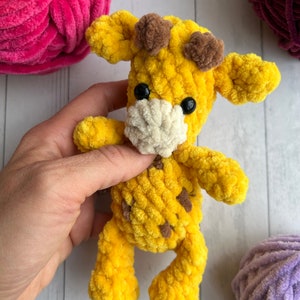 May include: A yellow crocheted giraffe toy with brown ears and black eyes. The giraffe is holding a white crocheted bib.