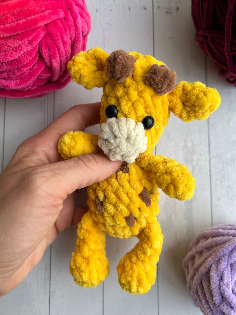 May include: A yellow crocheted giraffe toy with brown ears and black eyes. The giraffe is holding a white crocheted bib.