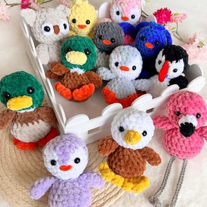 May include: A collection of handmade crochet animal plushies, including ducks, penguins, and an owl. The plushies feature various colors like green, yellow, pink, and blue. They are arranged in a white tray, with pink flowers in the background.