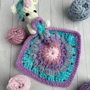 Op de afbeelding: Een gehaakt unicorn knuffeldoekje met een paars, blauw en roze kleurenschema. Het knuffeldoekje is gemaakt van zacht, pluche garen en heeft een gehaakt unicorn hoofdje bevestigd aan een vierkant deken.