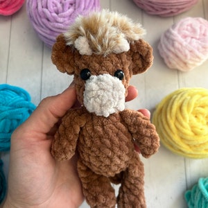 May include: A brown crocheted stuffed animal resembling a highland cow with a white face and brown fur. The cow has black eyes and a brown fuzzy mane. The cow is sitting on a hand.