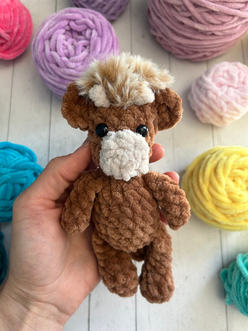 May include: A brown crocheted stuffed animal resembling a highland cow with a white face and brown fur. The cow has black eyes and a brown fuzzy mane. The cow is sitting on a hand.