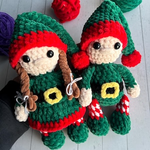 May include: Two crocheted elf dolls with green and red outfits. The dolls have brown yarn hair and black eyes.