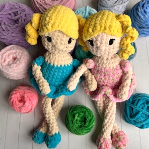 May include: Two crocheted dolls with yellow yarn hair and blue and pink dresses. The dolls are sitting on a white wooden surface with balls of yarn in various colors.
