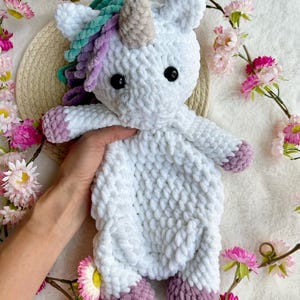 May include: A white crocheted unicorn toy with a multi-coloured mane and horn. The unicorn has black eyes and purple hooves. The plush is surrounded by pink flowers.