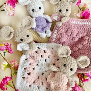 Pode incluir: Uma coleção de ratos de crochê feitos à mão em branco, cinza e rosa. Os ratos são colocados com cobertores de crochê rosa e branco e rodeados por flores rosa. Os brinquedos têm orelhas grandes e olhos pretos.