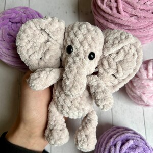 May include: A crocheted plush elephant toy in a light grey colour with black eyes. The elephant is sitting on a hand and surrounded by balls of yarn in pink and purple.