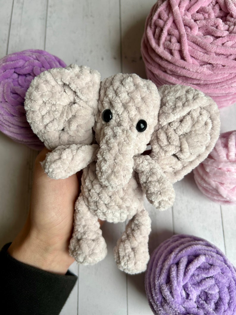 May include: A crocheted plush elephant toy in a light grey colour with black eyes. The elephant is sitting on a hand and surrounded by balls of yarn in pink and purple.