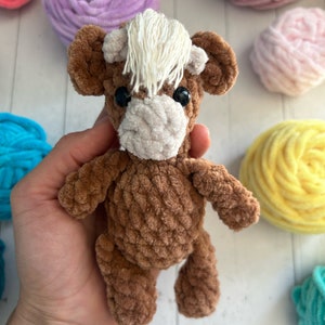 May include: A brown crocheted stuffed animal resembling a cow with white yarn for hair. The toy has black eyes and a white crocheted nose.
