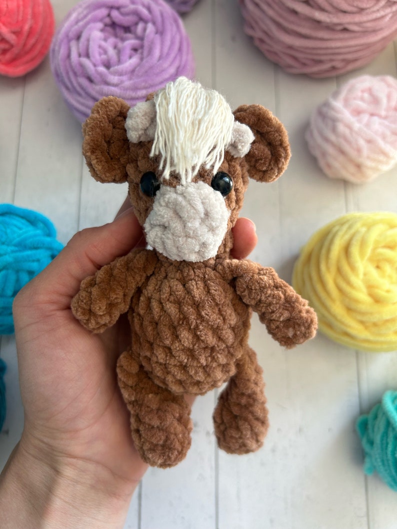 May include: A brown crocheted stuffed animal resembling a cow with white yarn for hair. The toy has black eyes and a white crocheted nose.