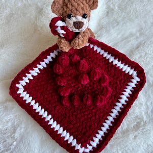 May include: A red and white crocheted baby blanket with a brown teddy bear holding a candy cane. The blanket has a textured design with small pom-poms.