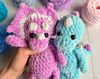 May include: Two crocheted dinosaur toys, one pink and one blue, with white accents. The toys are made with a soft, fuzzy yarn and have large, black eyes.