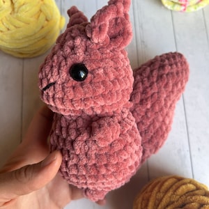 Peut inclure: Un jouet en peluche de l'écureuil crocheté de couleur rose poussiéreux avec des yeux noirs. L'écureuil est assis sur une surface blanche.