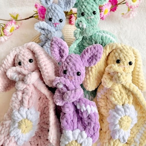May include: Four crocheted bunny baby blankets in pastel colours. Each blanket features a daisy design with a yellow centre and white petals. The bunnies are made with a soft, plush yarn.