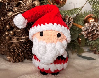 May include: A crocheted Santa Claus ornament with a red hat, white beard, and red and white striped suit. The ornament is sitting on a white fluffy surface.