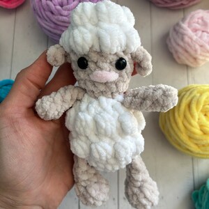 May include: A white crocheted sheep toy with black eyes and a pink nose. The sheep is sitting in a hand, surrounded by balls of yarn in various colours.