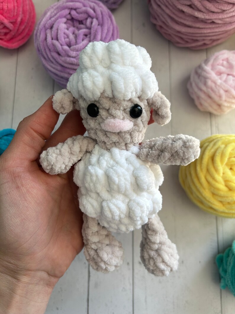 May include: A white crocheted sheep toy with black eyes and a pink nose. The sheep is sitting in a hand, surrounded by balls of yarn in various colours.