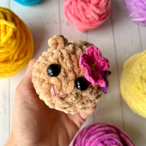 Puede incluir: Un animal de crochet marrón con grandes ojos negros y una flor rosa en la cabeza. El animal está siendo sostenido en una mano. Hay varias bolas de hilo en el fondo, incluyendo azul, rosa, amarillo y morado.