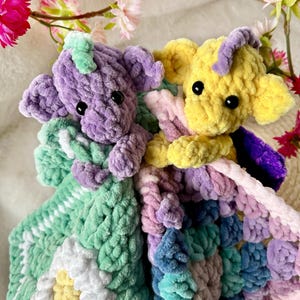 May include: Two handmade crochet stuffed animals, one purple and one yellow, each holding a colorful blanket. The blankets feature a daisy and a variety of pastel colors. The toys have black eyes and are surrounded by flowers.