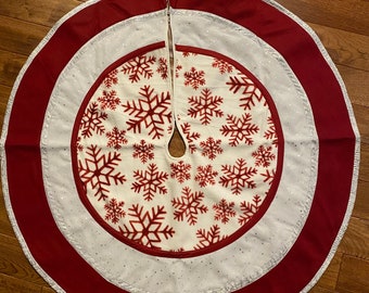 May include: Circular Christmas tree skirt with a white center featuring red snowflakes. The skirt has a red border and a silver trim. The design is festive and seasonal.