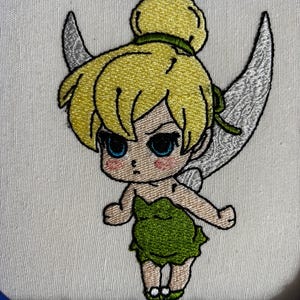 May include: Embroidered image of a cartoon fairy with yellow hair, green dress, and silver wings. The fairy has a stern expression with blue eyes and rosy cheeks. The embroidery is on a white fabric, framed in a blue hoop.