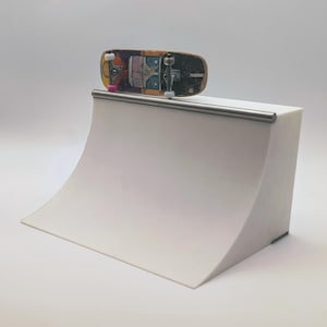 May include: A miniature skateboard with a colorful design rests on a white ramp with a silver rail. The skateboard features a worn, multi-colored deck and silver trucks. The ramp is a simple, curved structure.