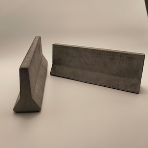 Puede incluir: Dos barreras Jersey en miniatura de hormigón gris. Las barreras son rectangulares con un frente inclinado y una parte superior plana. Están diseñadas para uso decorativo o como modelo.