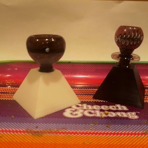 May include: Two glass smoking pipes on a colorful tray. The pipe on the left is brown and sits on a white pyramid-shaped base. The pipe on the right is purple with white designs and sits on a black pyramid-shaped base. The tray has a "Cheech & Chong" logo.