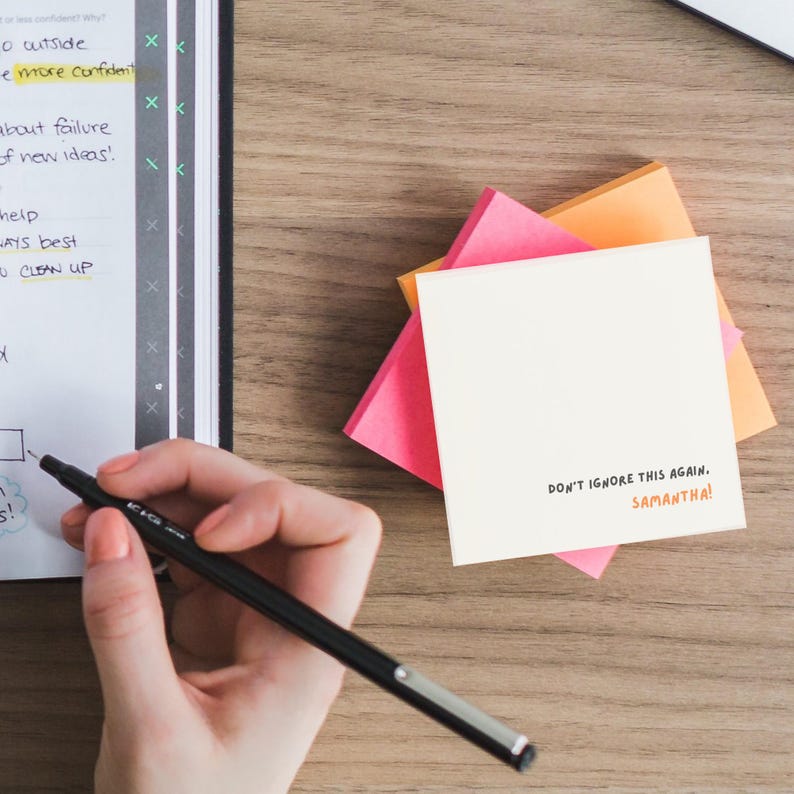 Op de afbeelding: Een witte sticky note met de tekst "DON'T IGNORE THIS AGAIN. SAMANTHA!" geschreven met een zwarte marker. De note is gestapeld op twee andere sticky notes, een roze en een oranje.