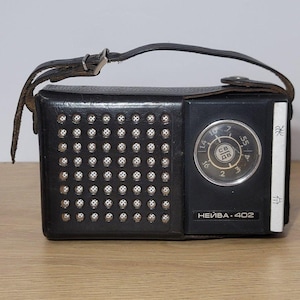Könnte beinhalten: Ein schwarzes Vintage-Radio mit Lederriemen. Das Radio hat einen Lautsprechergrill mit silberfarbenen Nieten und ein kreisförmiges Zifferblatt. Der Text "HEIBA 402" ist auf der Vorderseite aufgedruckt. Das Radio steht auf einer Holzoberfläche.