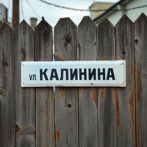 Puede incluir: Una valla de madera desgastada con una señal de calle rectangular blanca. La señal muestra el texto cirílico "ул. КАЛИНИНА" en letras negras. La valla está hecha de tablones de madera verticales con un aspecto gris envejecido.
