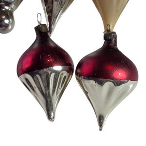 May include: A collection of glass Christmas ornaments featuring a teardrop shape with a pointed base. The ornaments have a two-tone design with a deep red matte upper section and a reflective silver lower section with vertical ribbed detailing.