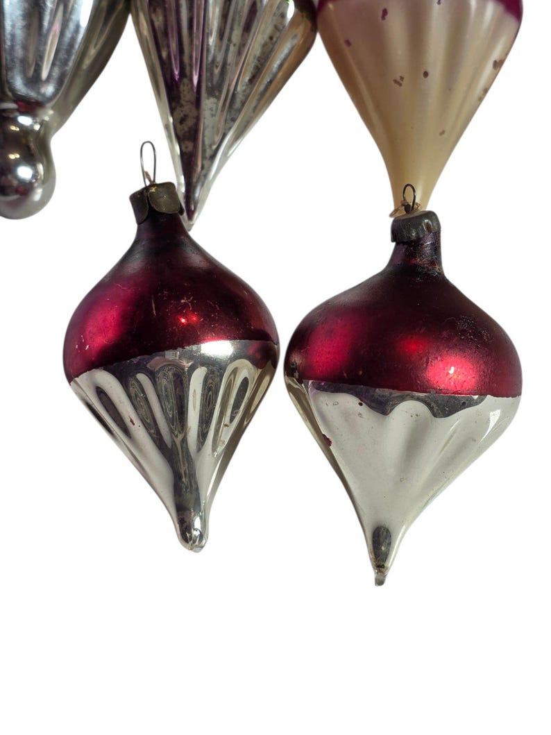 May include: A collection of glass Christmas ornaments featuring a teardrop shape with a pointed base. The ornaments have a two-tone design with a deep red matte upper section and a reflective silver lower section with vertical ribbed detailing.