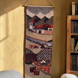 May include: A rectangular woven wall hanging featuring a landscape scene with mountains, white buildings with red roofs, and figures in traditional clothing. The textile art displays a color palette of tan, brown, red, blue, and white threads.