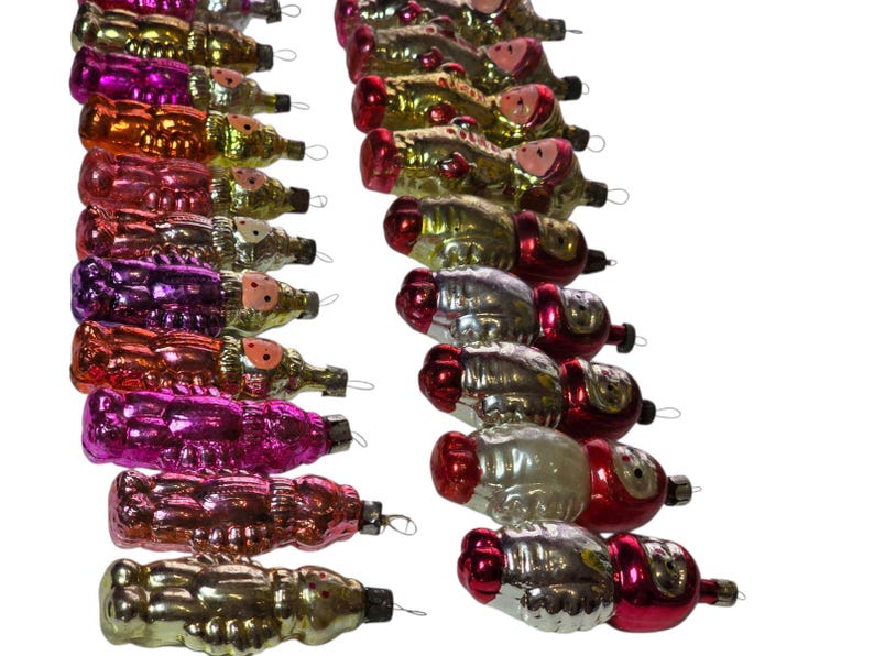 May include: A collection of vintage Christmas ornaments in various colors, including gold, pink, red, and purple. The ornaments are shaped like Santa Claus figures and have small metal loops for hanging. The ornaments are arranged in rows against a white background.