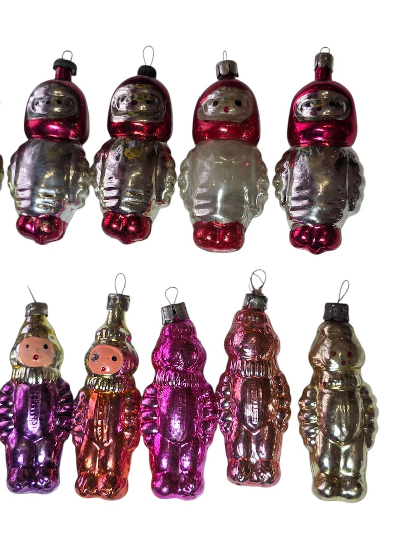 May include: A collection of vintage Christmas ornaments. The ornaments are shaped like figures, with a mix of colors including silver, red, pink, and gold. Each ornament has a loop for hanging.