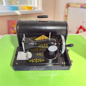 Könnte beinhalten: Eine schwarze Spielzeug-Nähmaschine im Vintage-Stil mit passendem schwarzem Tragekoffer. Die Maschine ist mit goldenen und roten floralen Rankenmustern, einer silbernen Stichplatte, einem Handkurbelrad und einem kleinen gelben Rauten-Emblem verziert.