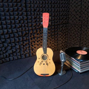 Può includere: Una chitarra acustica in legno chiaro con paletta e ponte arancione brillante. Il corpo presenta disegni a linee nere di due uccelli e una stella attorno alla buca. Un microfono argentato in stile vintage si trova accanto a dei dischi in vinile.
