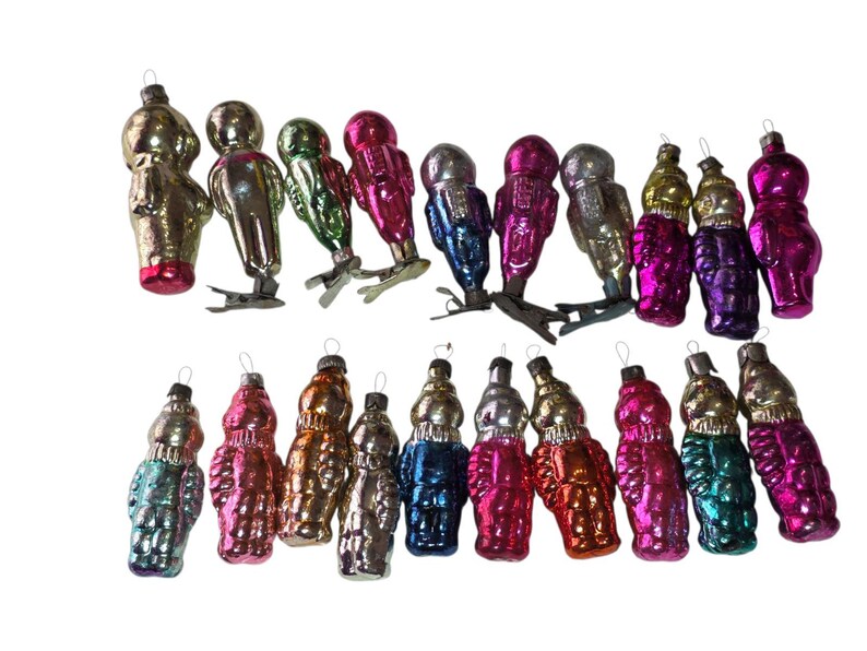 May include: A collection of twenty vintage glass ornaments shaped like figures, featuring a variety of metallic and glossy finishes in colors including pink, gold, blue, green, and silver. Several ornaments include metal clips at the base.