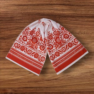 May include: White and red floral patterned scarf. The scarf features a repeating floral design in red against a white background. The edges are trimmed with a red border and text. The scarf is folded and displayed on a wooden surface.
