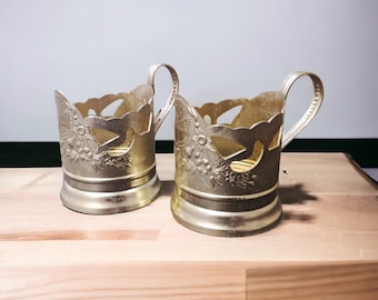 May include: Two ornate silver-toned metal cup holders with handles. The holders feature floral and leaf designs with cut-out patterns. They sit on a light-colored wooden surface, with a plain white background.