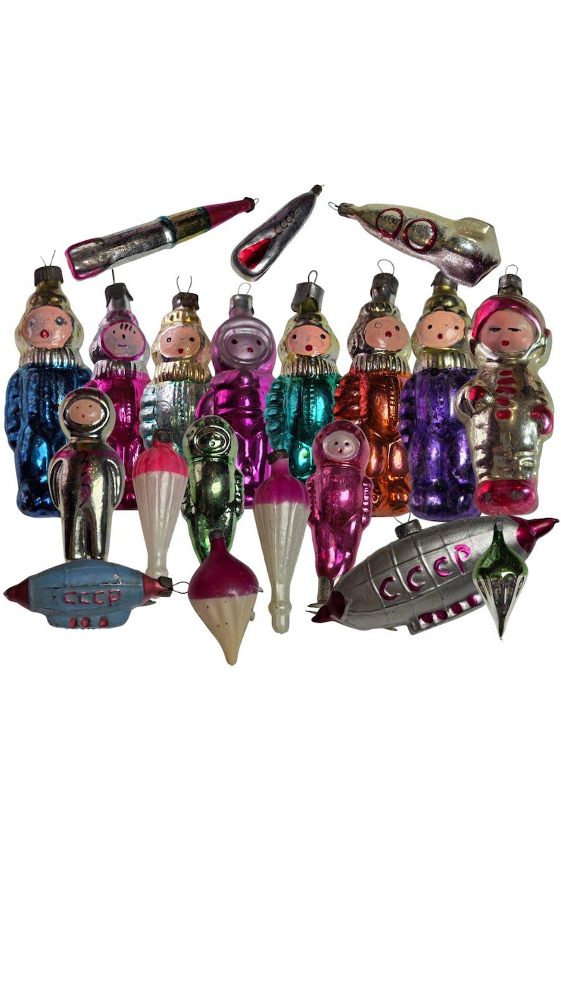 May include: A collection of vintage glass Christmas ornaments featuring various figures, including cosmonauts in colorful suits, rockets, and airships. Several pieces are marked with the Cyrillic text CCCP, representing Soviet-era space themes.