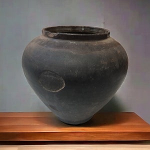 Puede incluir: Una olla grande de cerámica gris oscuro con una base redondeada y una abertura ancha. La olla está sentada sobre una plataforma de madera.