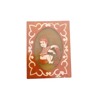 May include: A rectangular orange plaque featuring a central oval window with a graphic illustration of a child wearing a striped shirt, sitting and pouring liquid from a container. The border is decorated with a white, swirling, ornate pattern.