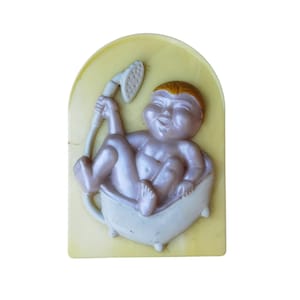 May include: A molded plastic wall plaque featuring a light purple baby with orange hair sitting in a white bathtub. The baby holds a white shower head attachment. The entire figure is set against a pale yellow, arched background panel.